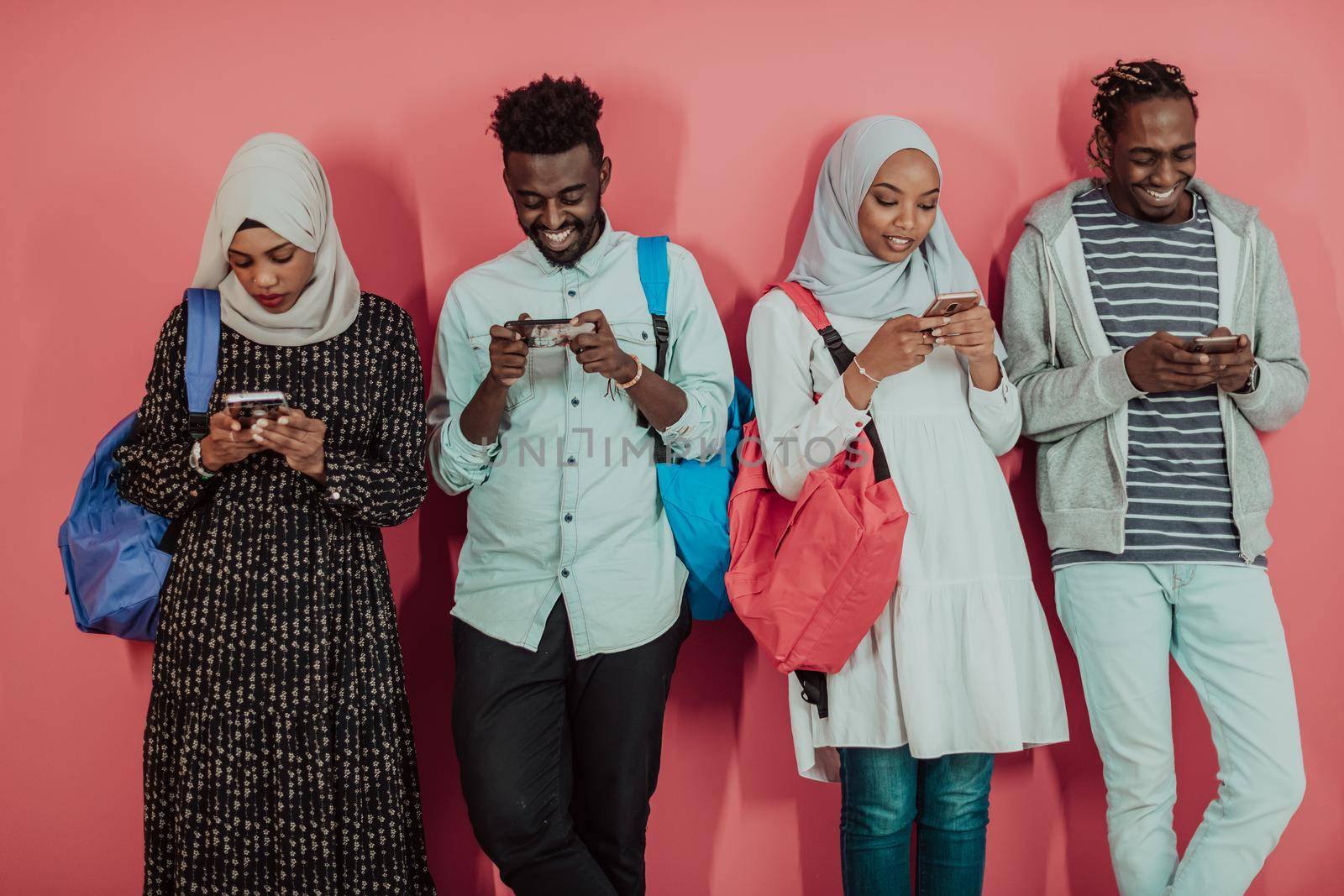 A group of African Muslim students use smartphones while standing in front of a pink background by dotshock