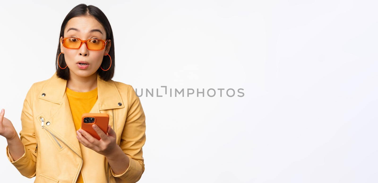Portrait of korean girl in sunglasses, holding smartphone, looking confused and shrugging, standing over white background. Copy space