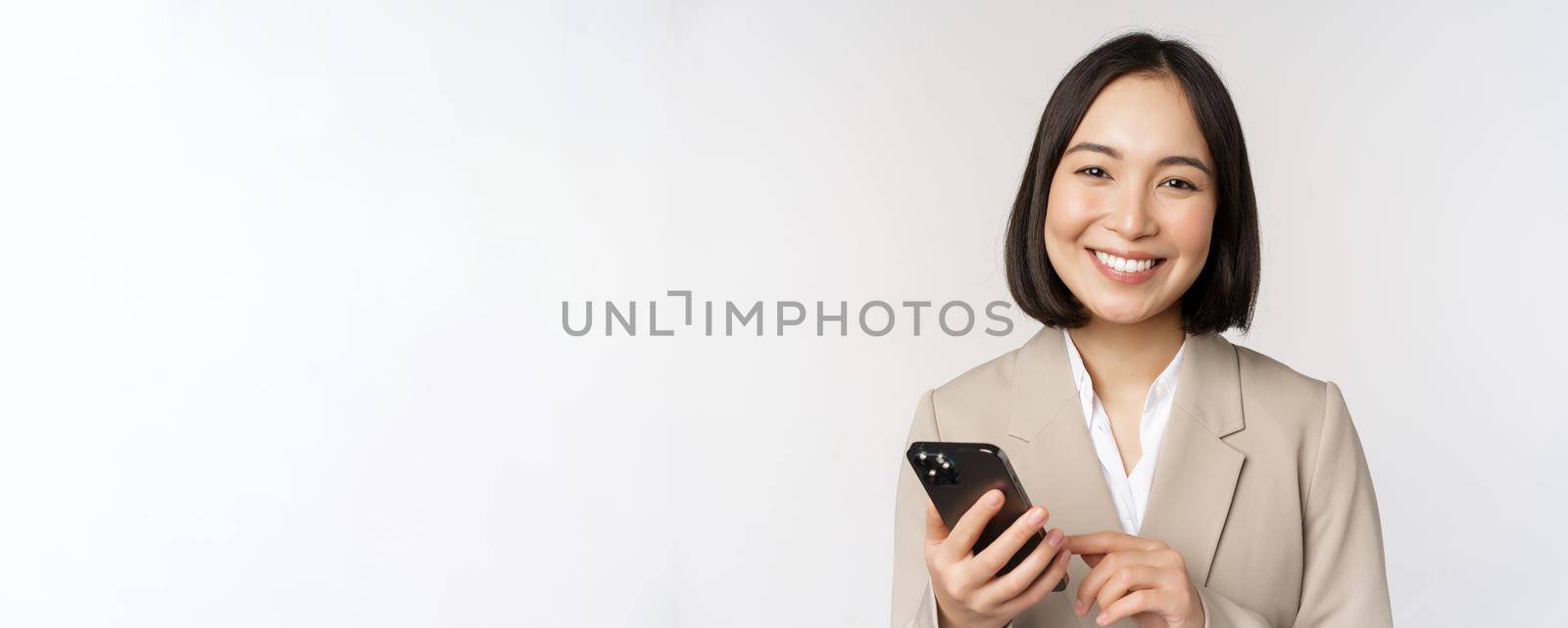 Close up portrait of korean woman, corporate lady in suit, using mobile phone and smiling, holding smartphone, standing over white background.