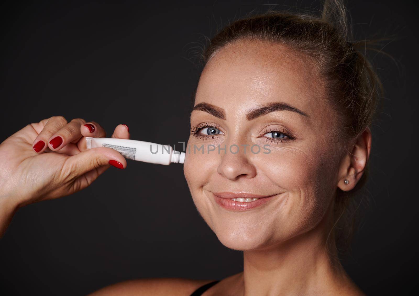 Beautiful blonde Caucasian woman applying anti wrinkles smoothing anti-aging under eyes serum or cream, caring of her face skin. Close-up, studio shot by artgf