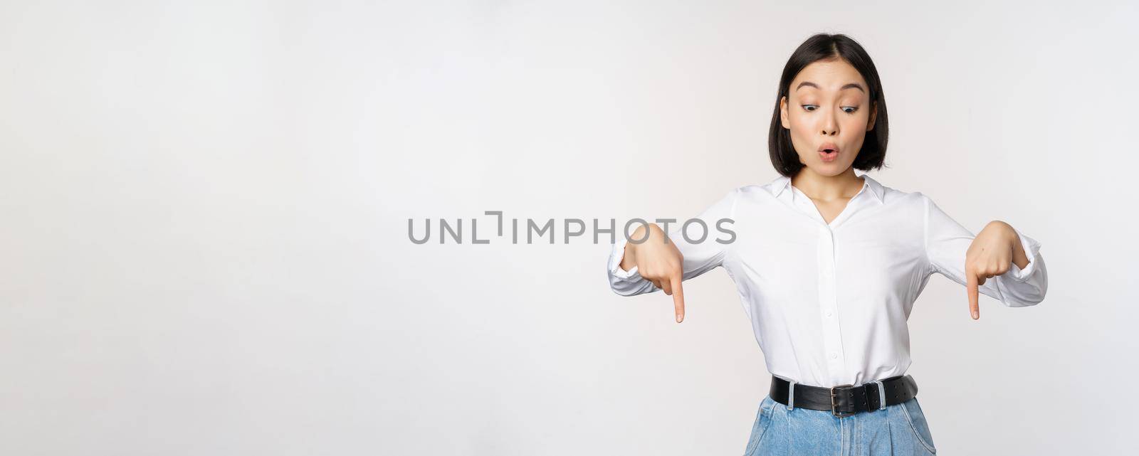 Surprised young asian female student, pointing fingers down and looking with amazed, impressed face expression, standing over white background.