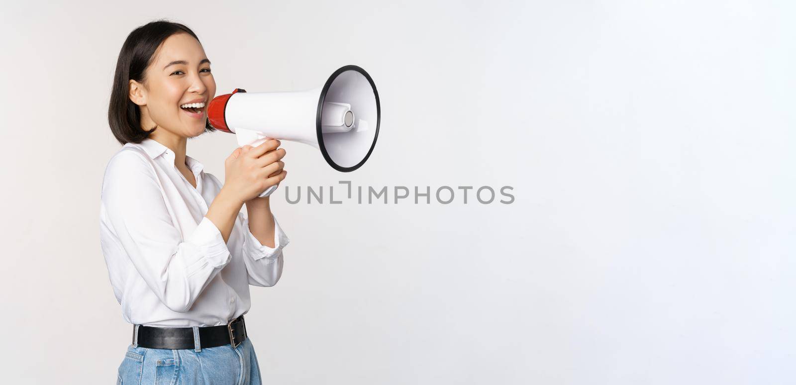 Image of young woman, korean activist, recruiter screaming in megaphone, searching, shouting at loudspeaker, standing over white background.