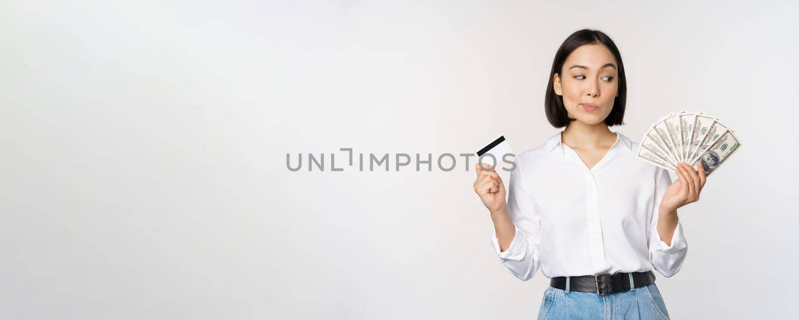 Image of asian woman looking at money dollars, holding credit card in another hand, thinking, standing over white background.