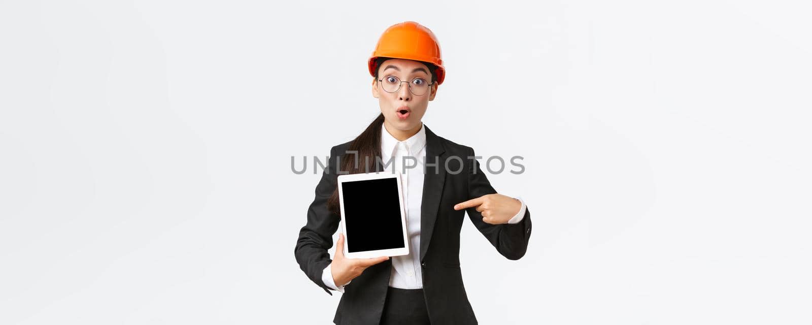 Impressed and surprised asian female engineer, technician at industrial area wear safety helmet and business suit, pointing finger at profit chart, diagram at digital tablet screen, look amazed.