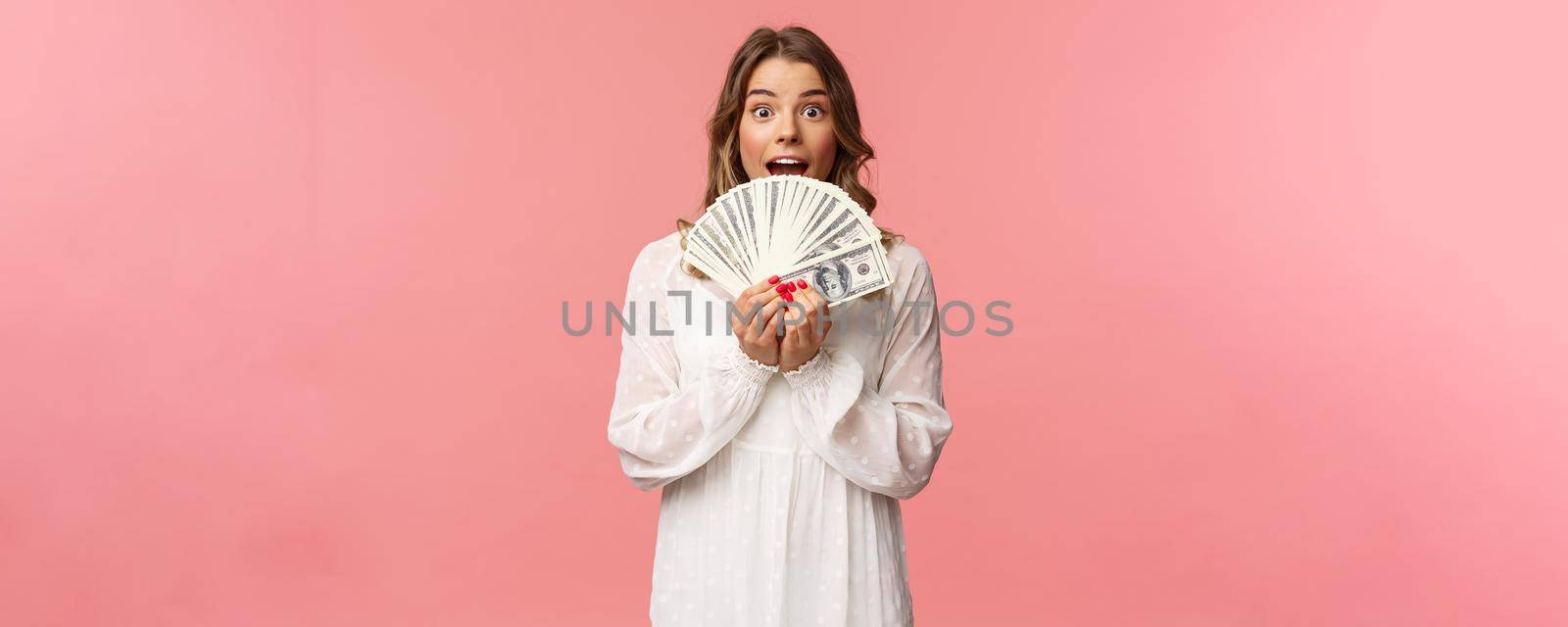 Portrait of excited lucky girl with blond hair, white dress, winning money, receive cash award, big lottery prize, holding dollars near face, smiling and looking amazed, standing pink background by Benzoix