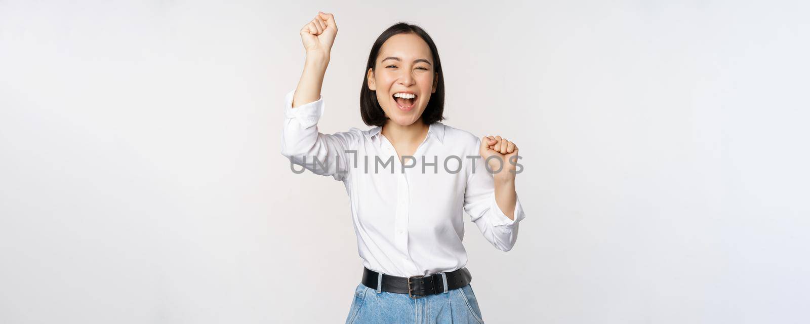 Image of happy lucky asian woman hooray gesture, winning and celebrating, triumphing, raising hadns up and laughing, standing over white background.