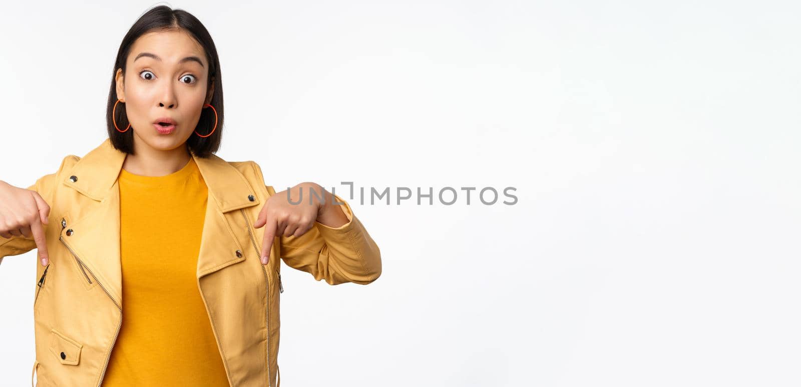 Portrait of surprised asian girl, looking amazed and impressed, pointing fingers down, showing banner with shocked face expression, white background. Copy space