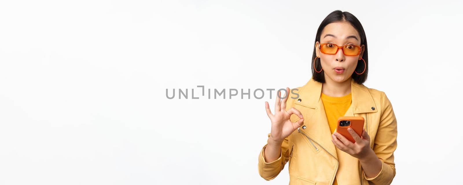 Amazed asian girl in sunglasses, showing okay, ok sign, holding smartphone, looking impressed, recommending smth, standing over white background.