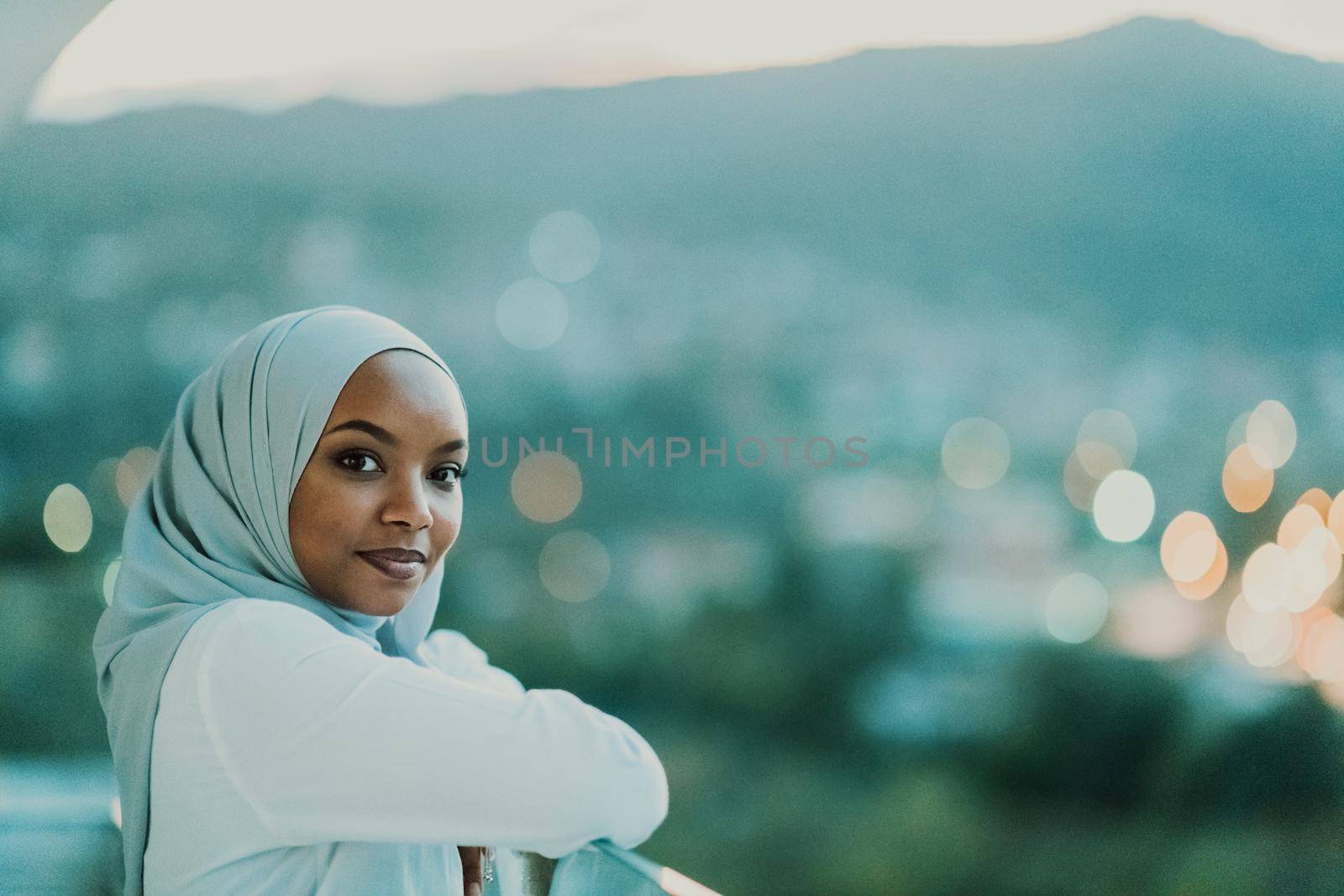 African Muslim woman in the night on a balcony smiling at the camera with city bokeh lights in the background. High-quality photo