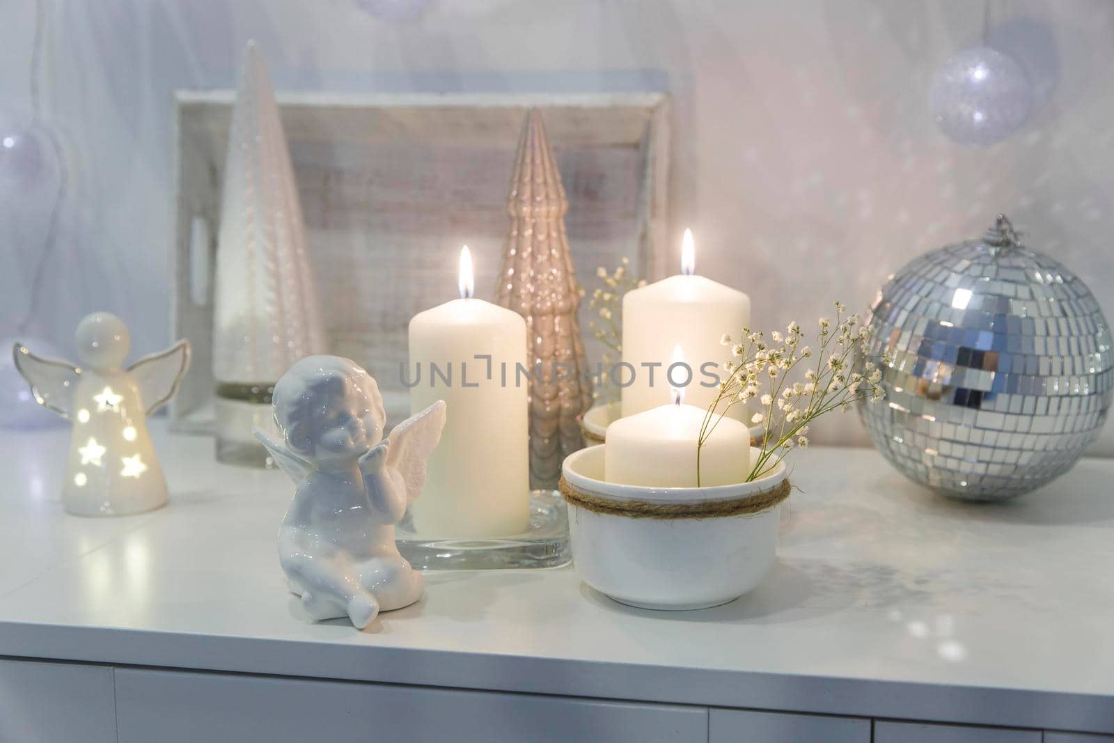 The concept of decorating the house for Christmas. Lighted candles, a garland, a disco ball, an angel figurine on a white chest of drawers. Place for text by elenarostunova