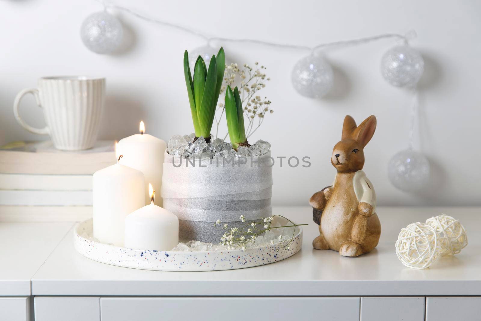 Unblown hyacinths with burning candles on a tray, a stack of books, a mug of tea, a garland of thread on the wall. Easter room decoration.