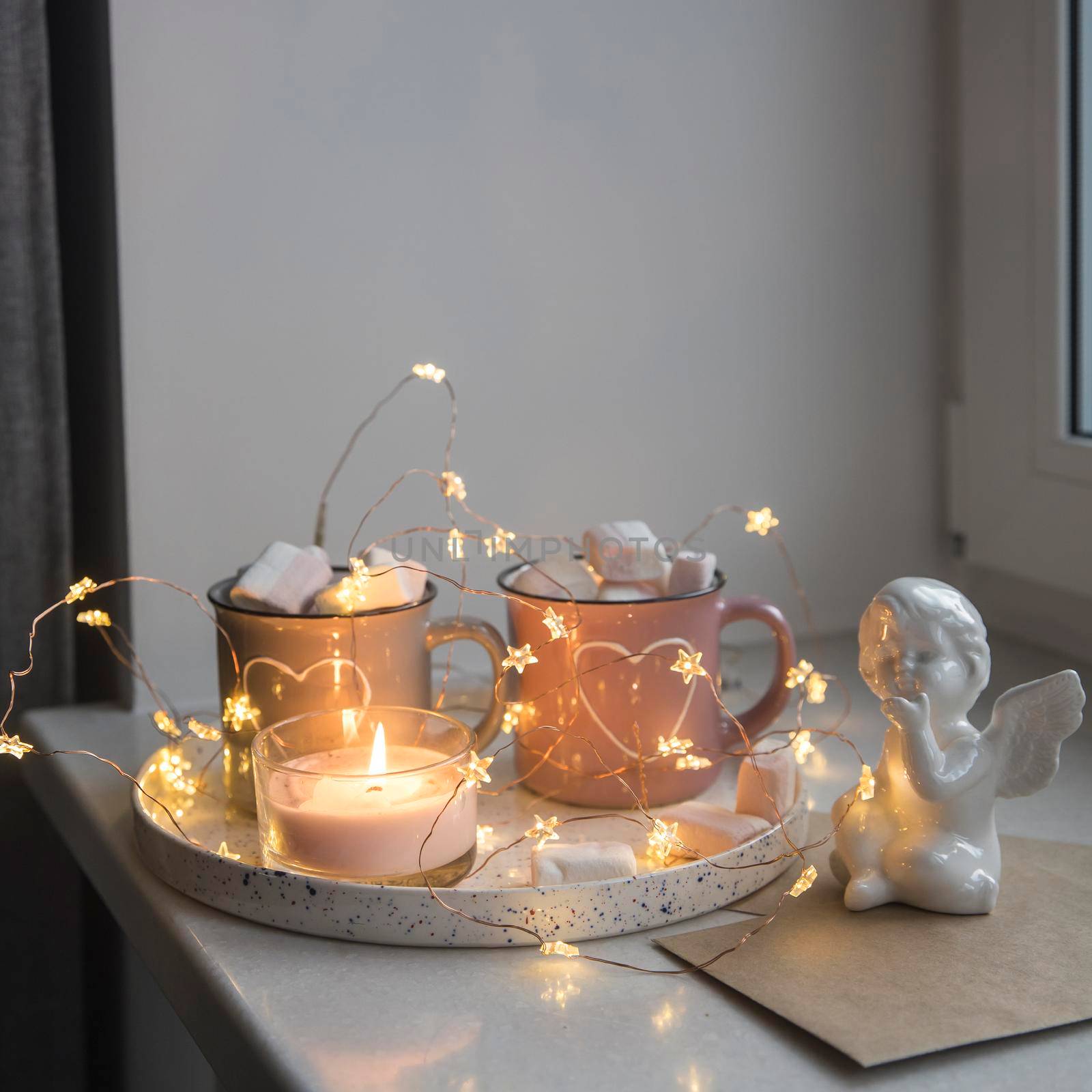Apartment decoration for Christmas. Gray and pink mugs with painted white hearts with cocoa, marshmallows on tray on the window, composition decorates the interior on the eve of Christmas. Copy space