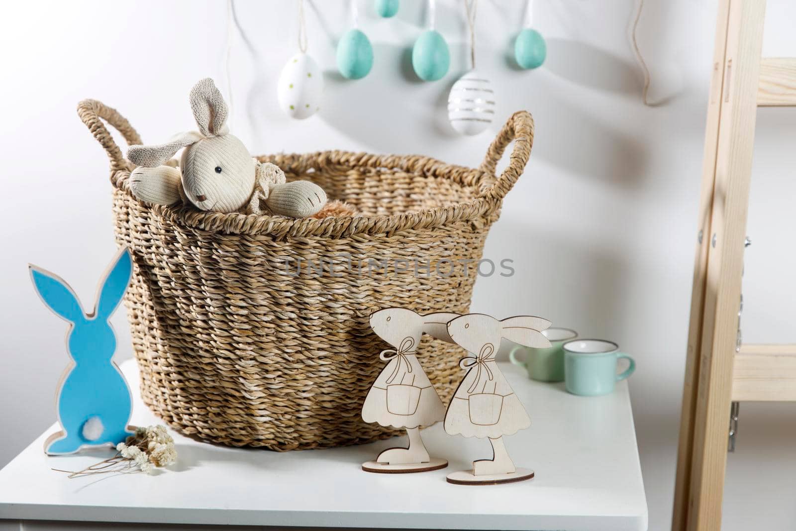 Interior decoration for Easter. A rag rabbit peeks out of a wicker willow basket on the table. A garland of wooden painted eggs above it.