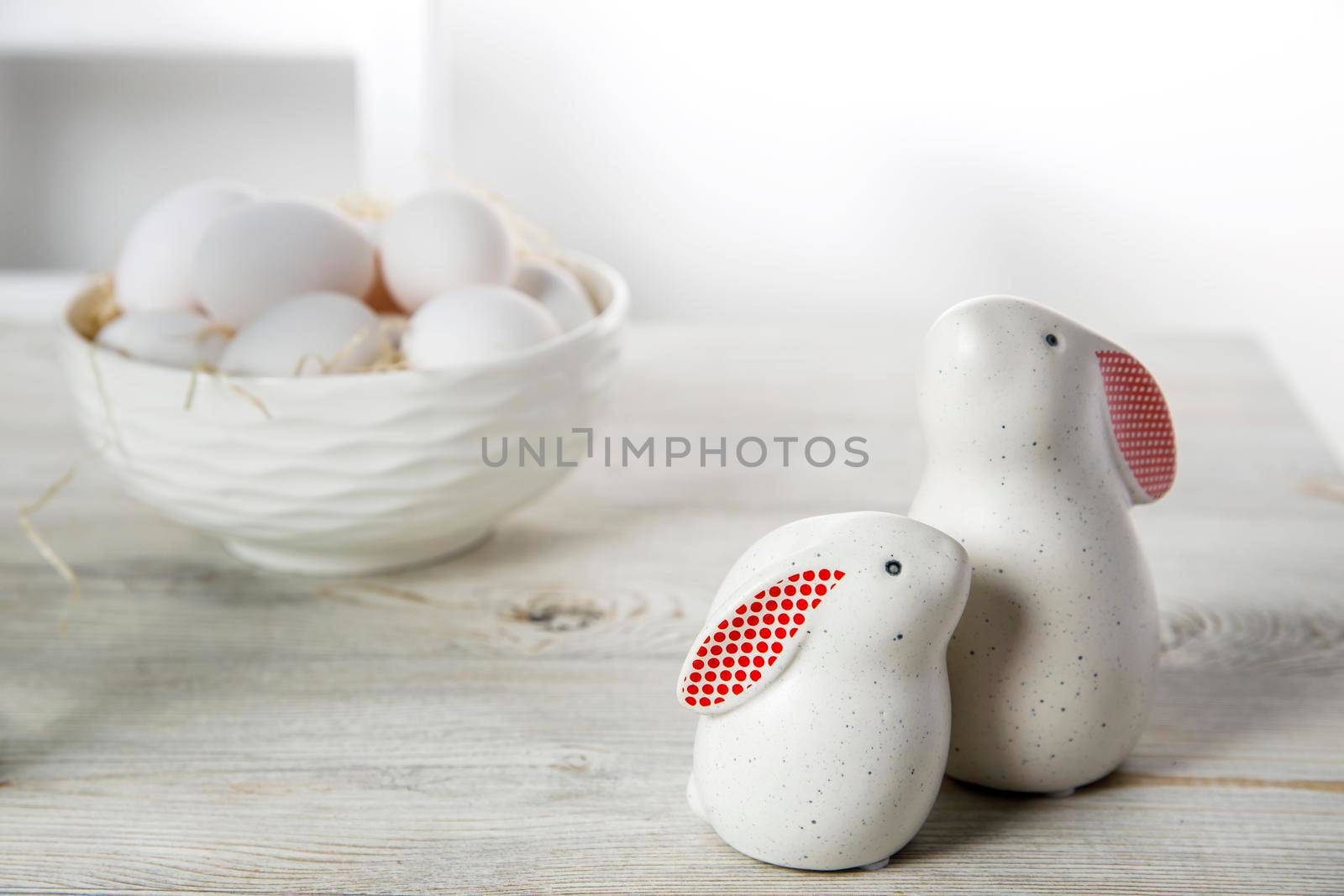 Two figurines of white hares with red ears big and small on a wooden saw cut and a bowl with eggs in the background in a white Scandinavian style kitchen