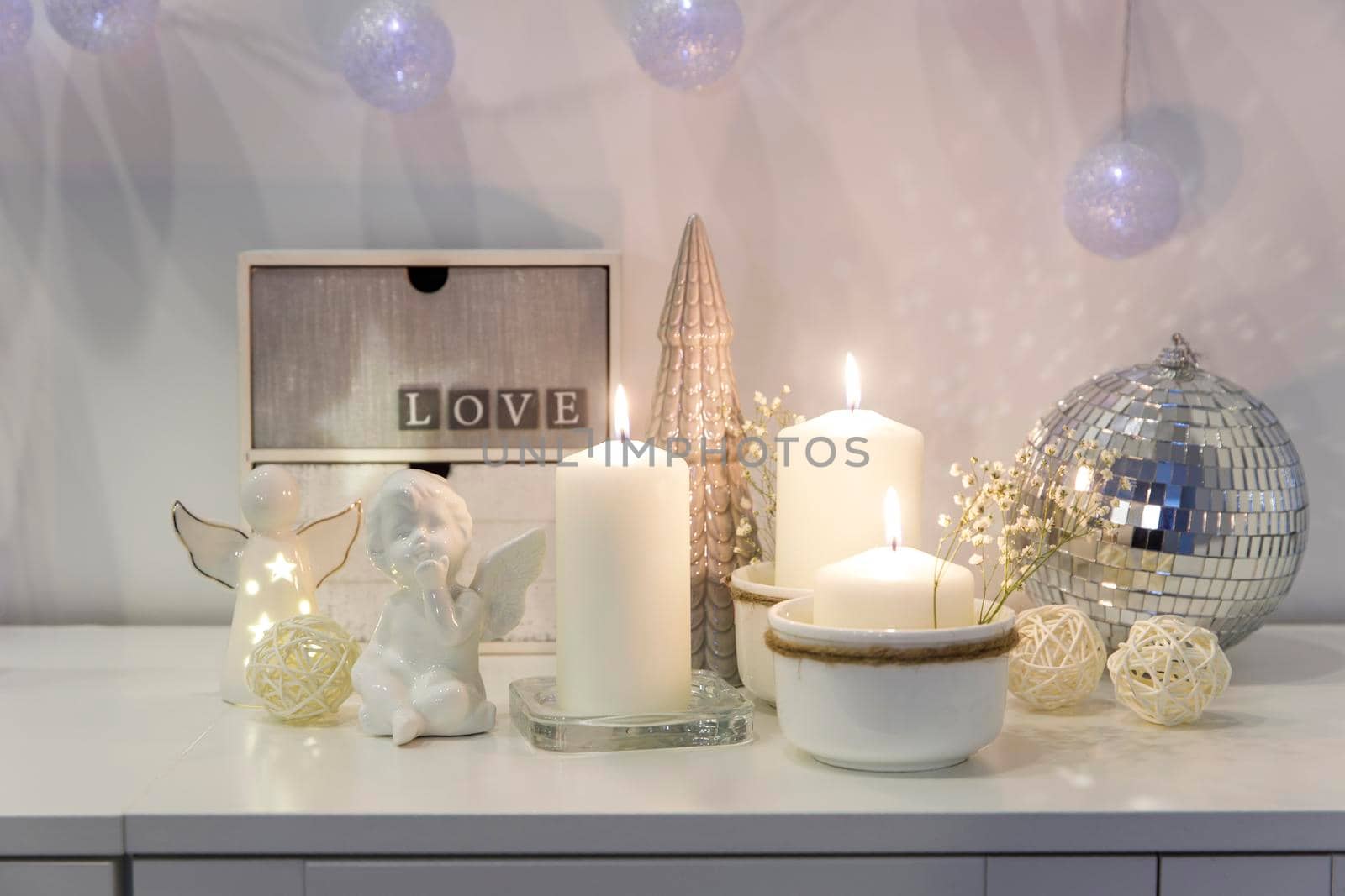 The concept of decorating the house for Christmas. Lighted candles, a garland, a disco ball, an angel figurine on a white chest of drawers. Place for text