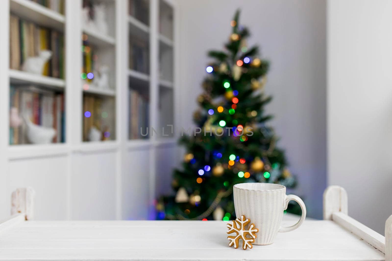 White cup with tea and gingerbread cookies in the form of a snowflake on the table against the background of an unsharp Christmas tree with lights in the interior