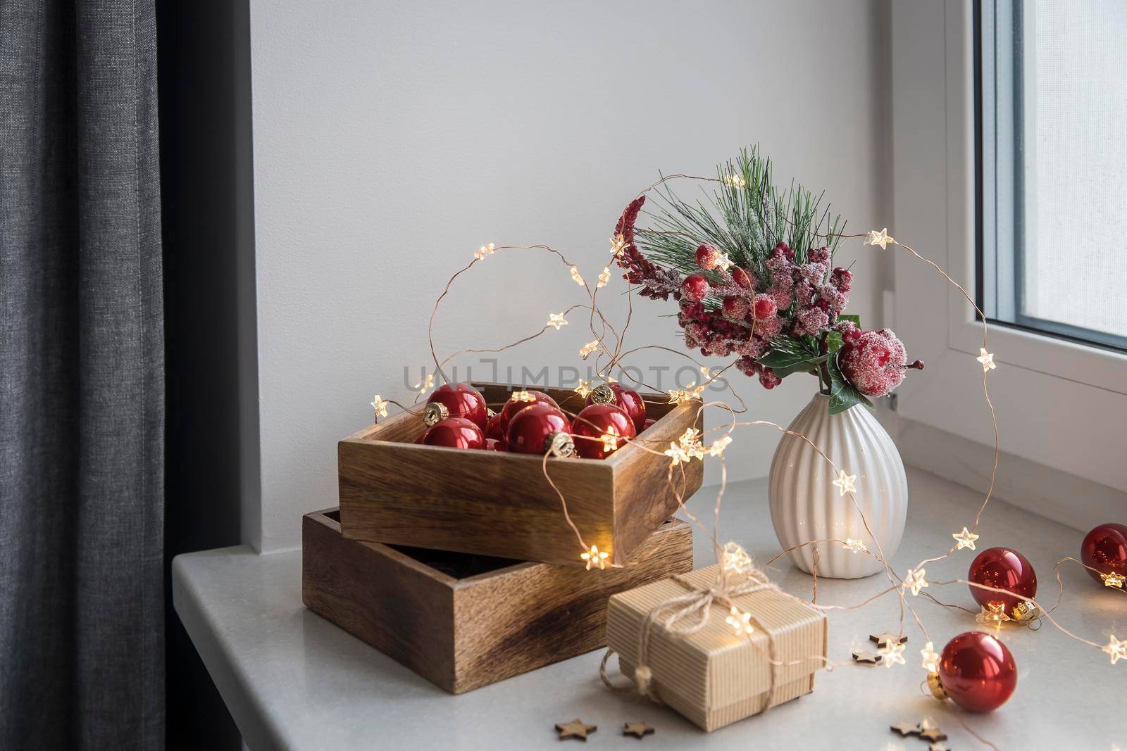 An artificial spruce branch, decorated with ivy leaves, apples and hawthorn berries. Lighted candles in wooden box, red glass balls, winter composition decorates interior on eve of Christmas by elenarostunova