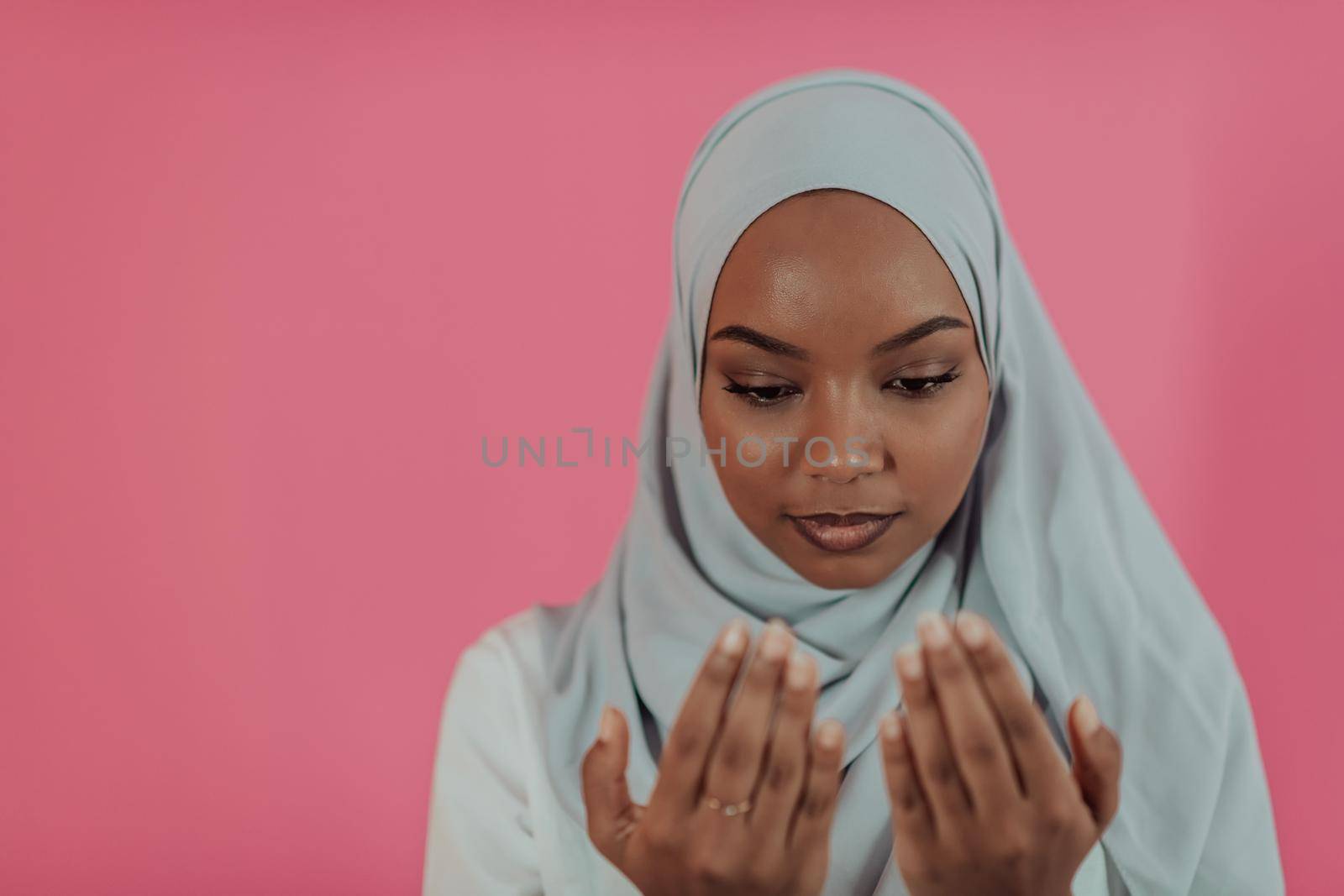 Modern African Muslim woman makes traditional prayer to God, keeps hands in praying gesture, wears traditional white clothes, has serious facial expression, isolated over plastic pink background. High-quality photo