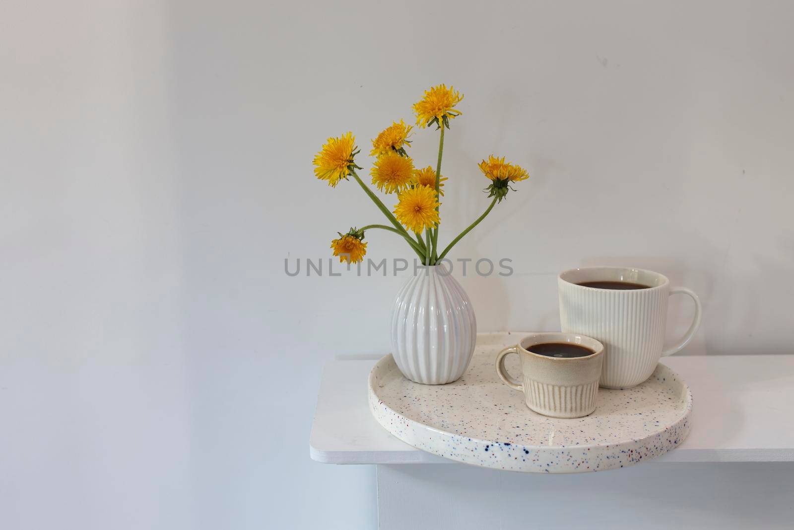 A bouquet of dandelions in a white fluted vase and two cups of different sizes with coffee on a white table. Breakfast. Copy space