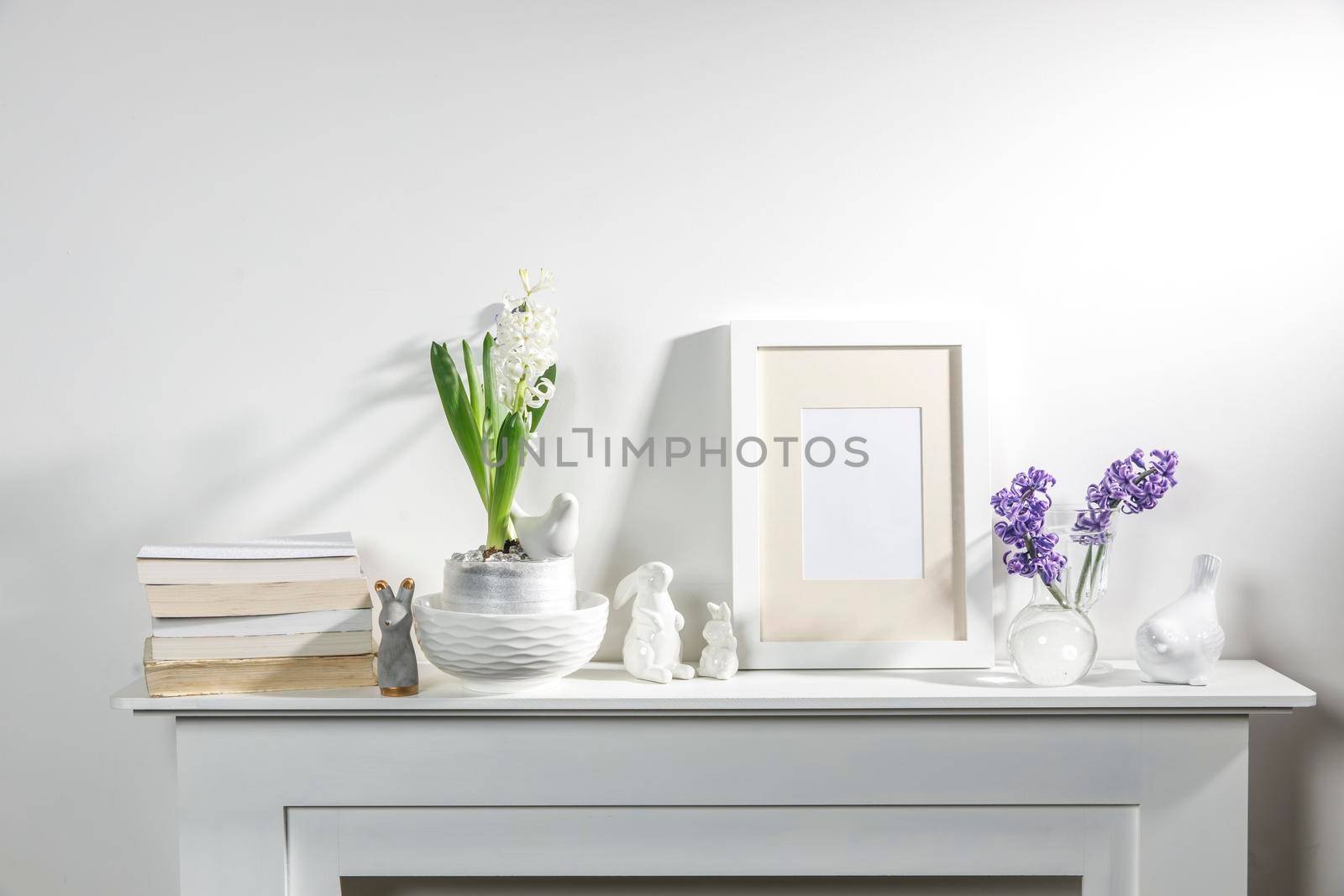 White hyacinth in a large porcelain bowl, books, figurines of hares and a bird, an empty photo frame are on the fireplace against the white wall. Layout. by elenarostunova