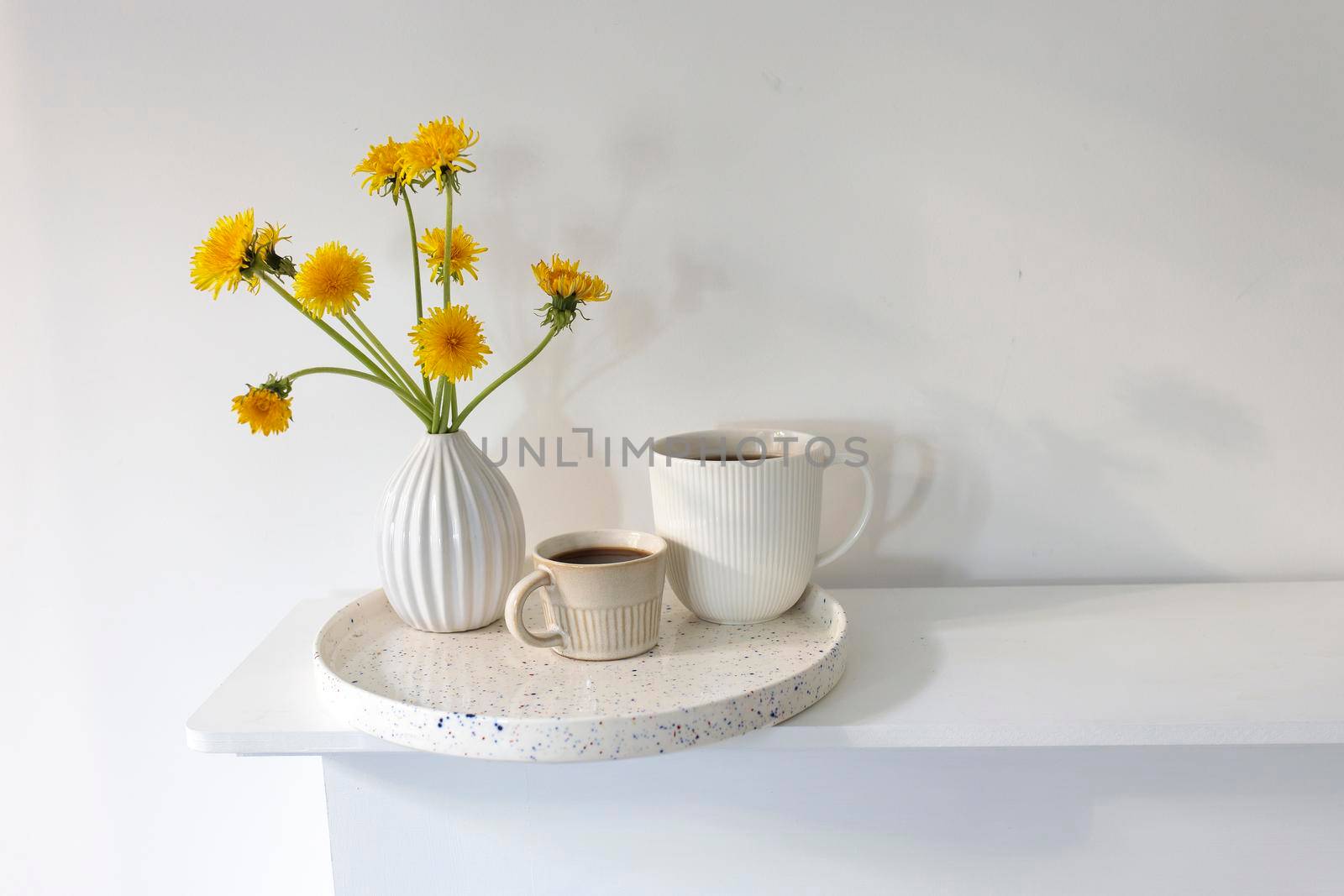 A bouquet of dandelions in a white fluted vase and two cups of different sizes with coffee on a white table. by elenarostunova