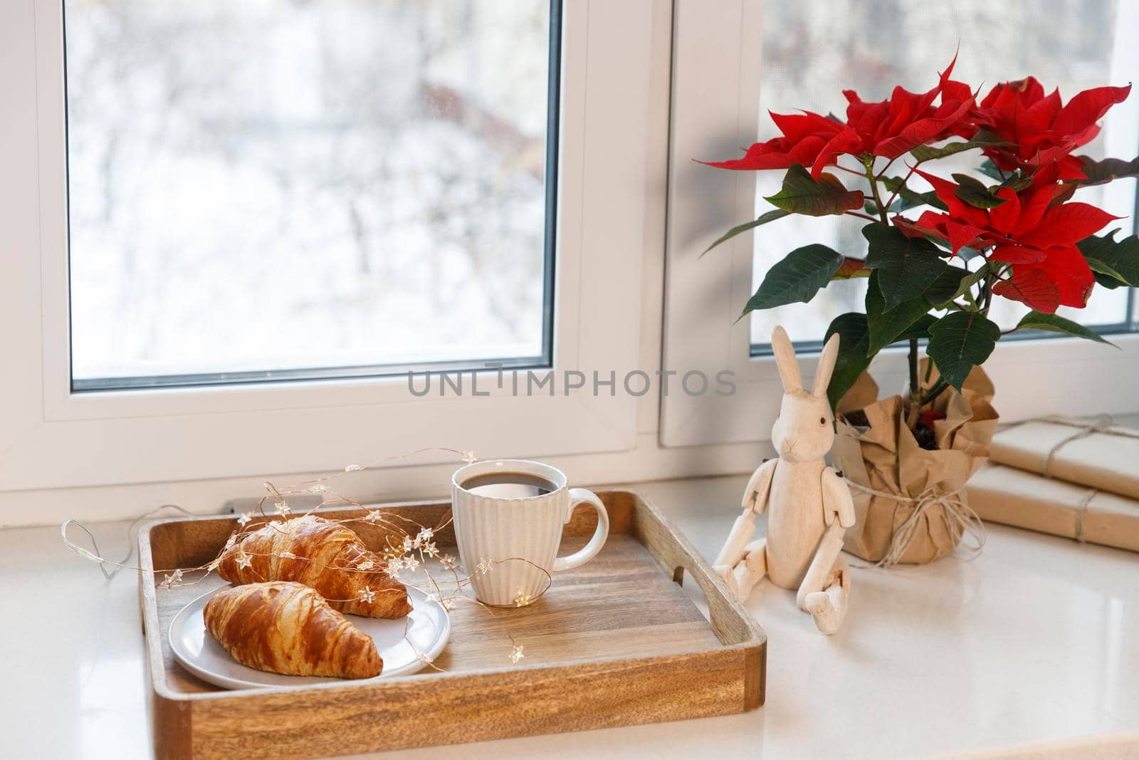 Freshly baked croissant on a gray round plate, a white cup of coffee and a garland on a tray on the windowsill. Blooming poinsettia and hinged wooden hare. by elenarostunova