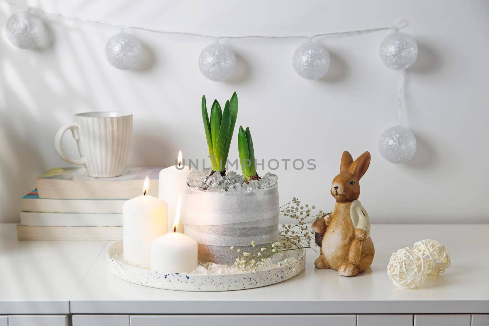 Unblown hyacinths with burning candles on a tray, a stack of books, a mug of tea, a garland of thread on the wall. Easter room decoration. by elenarostunova