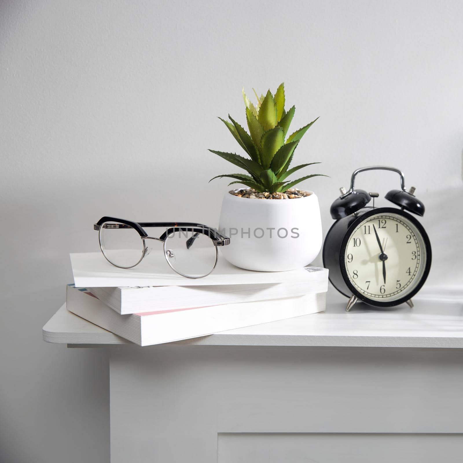 A stack of books, an artificial succulent plant in a ceramic pot, glasses, a metal basket with notebooks, a pen and a cup on a white table surface. Concept - office at home. Online training.