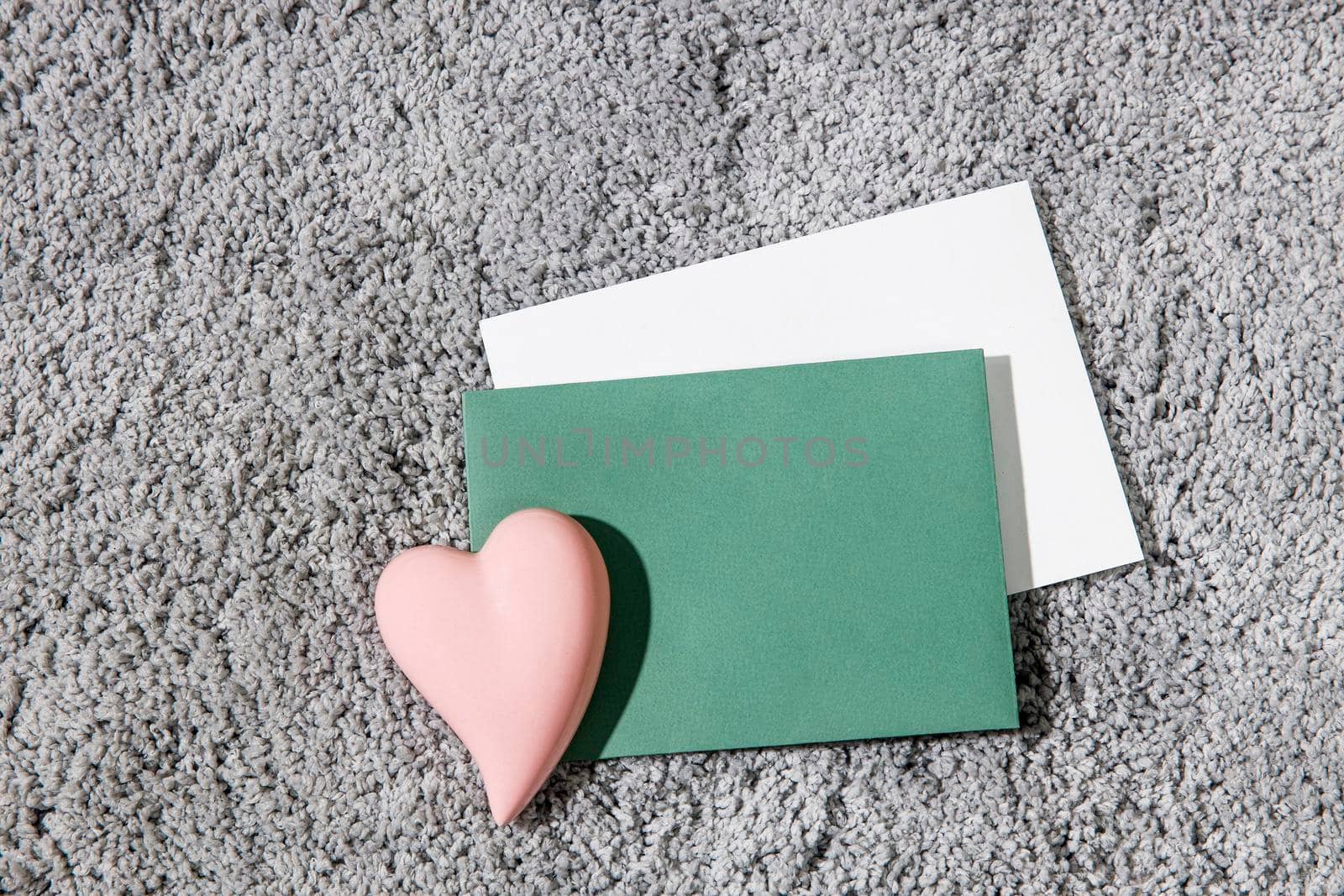 Greeting card for Valentine's Day. White and green envelopes, ceramic pink heart on a grey fluffy carpet background. Copy space. Place for text