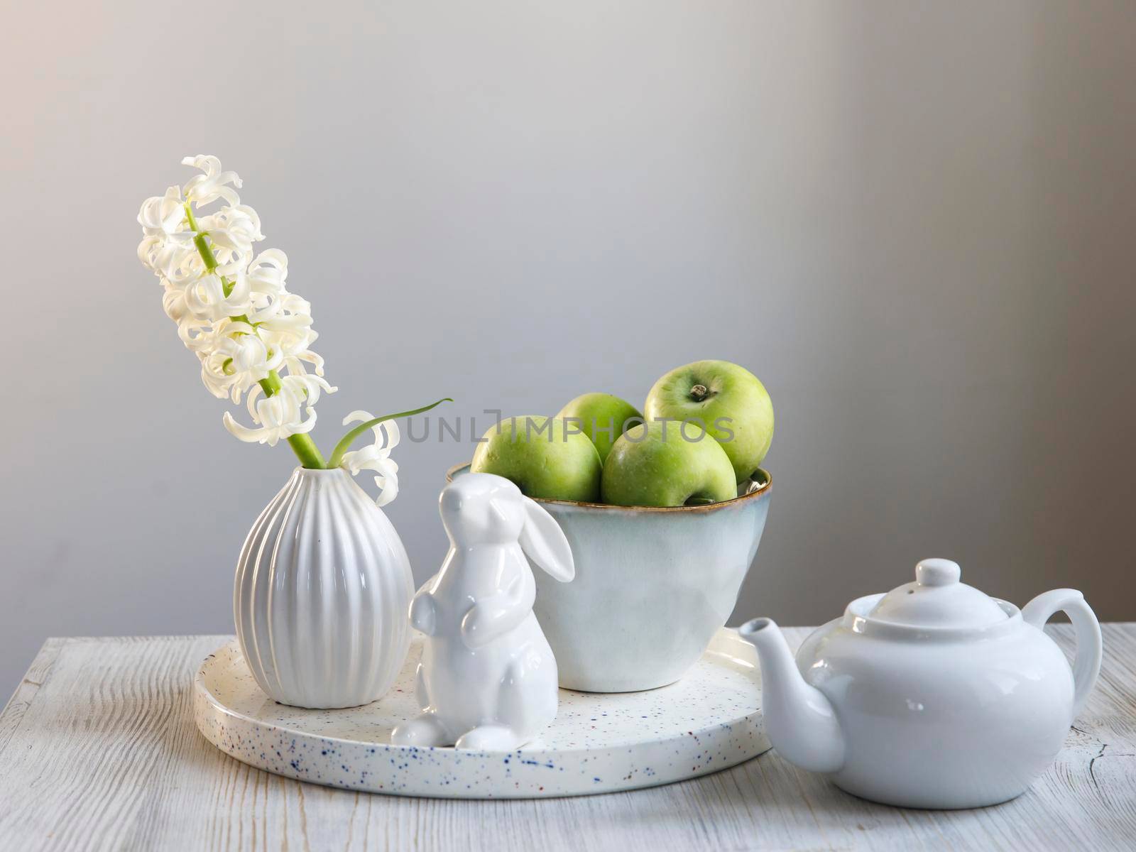 A white hyacinth in a 70s-style fluted vase next to a tray with a large bowl of green apples, an Easter china bunny and a teapot is on the table. Minimalism. Scandinavian style. by elenarostunova
