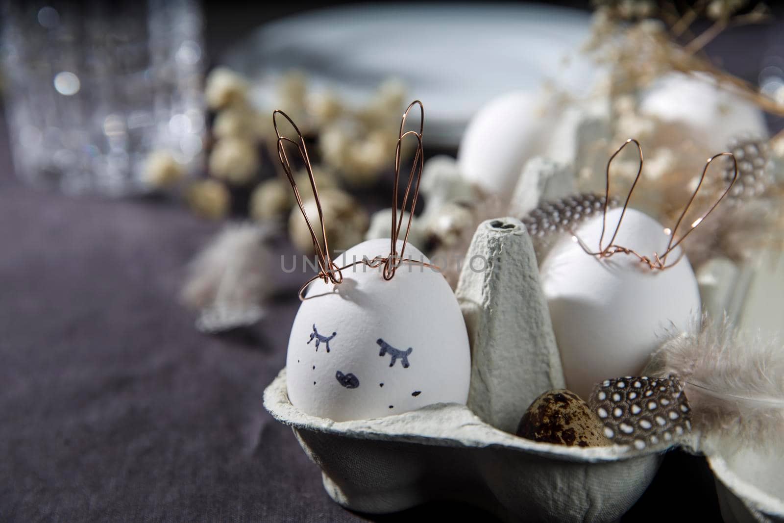 Easter concept. A white egg with copper wire ears and a painted rabbit sleeping face in a box. Partridge eggs and feathers. by elenarostunova