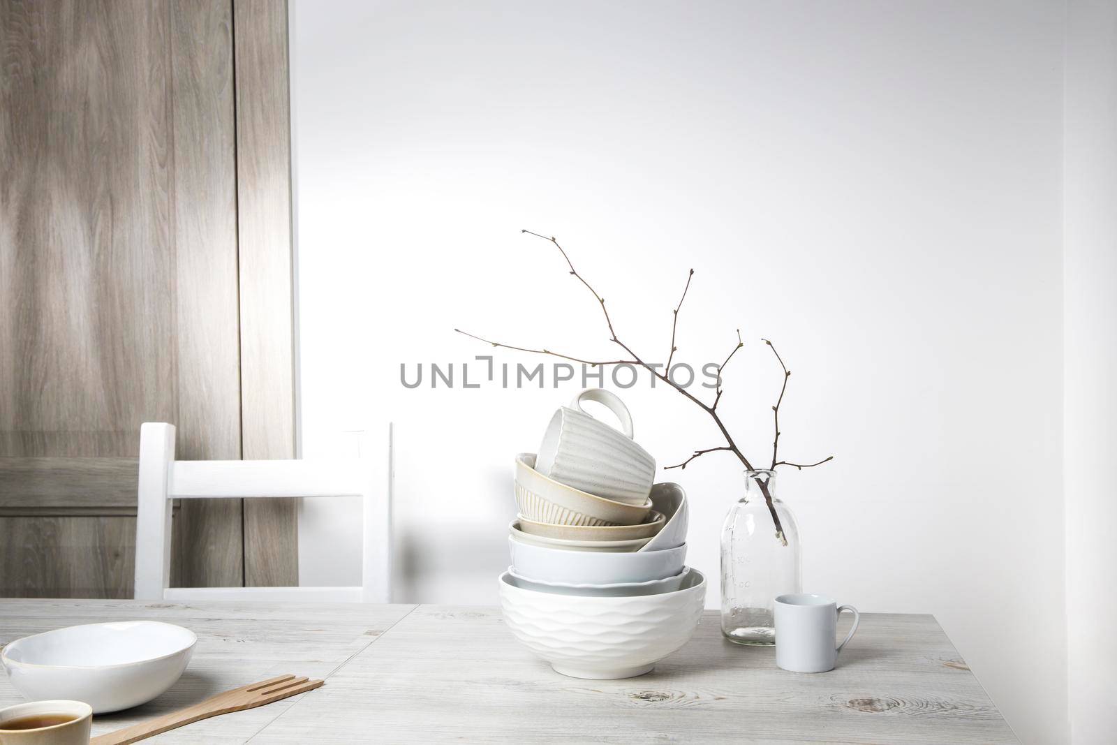 A pyramid of white bowls, plates and cups of different sizes and colors on a beige table in the kitchen. An unblown linden branch in a glass vase. Scandinavian style. Place for your text