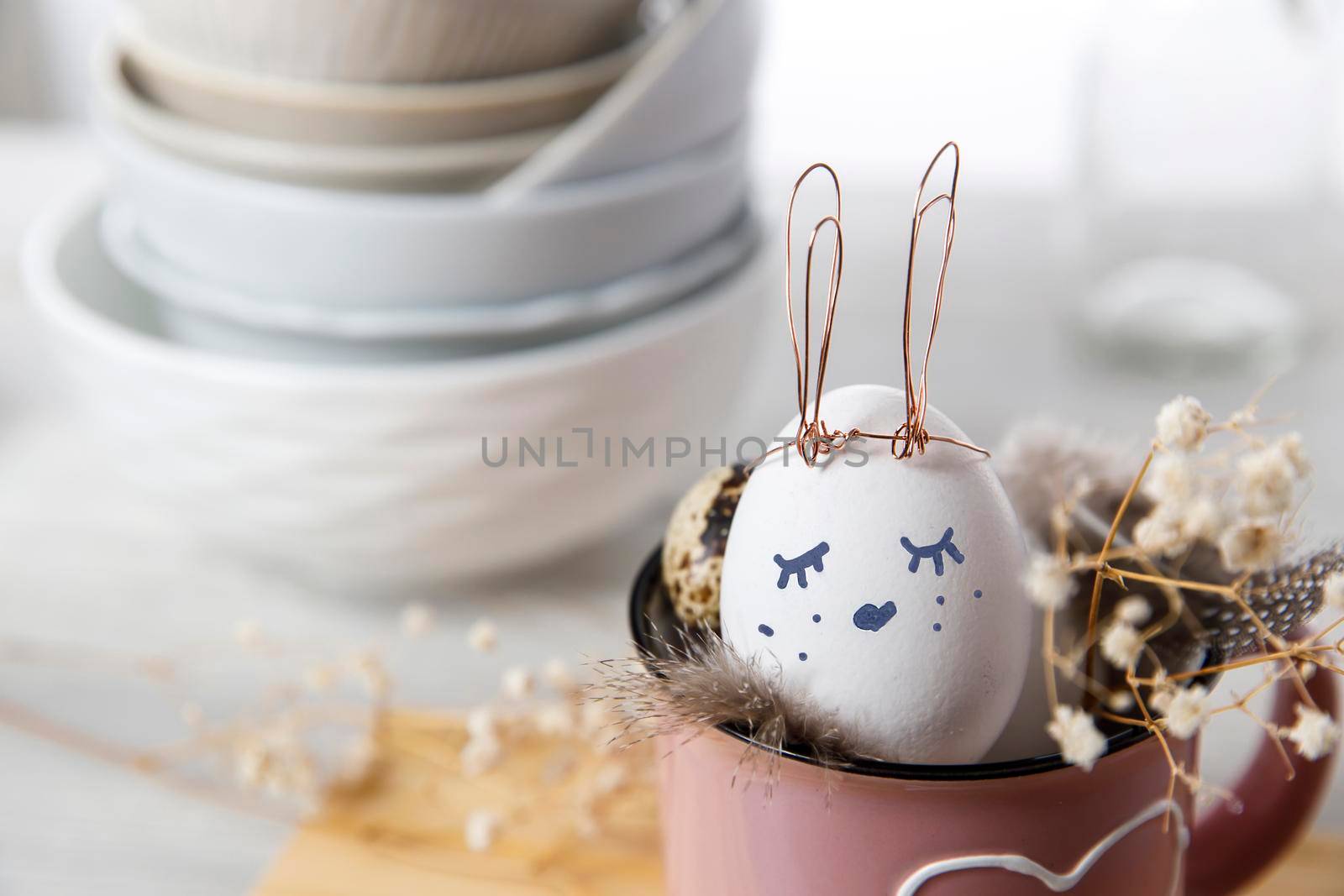 Easter concept. A white egg with copper wire ears and a painted rabbit sleeping face in a cup. Partridge eggs and feathers.