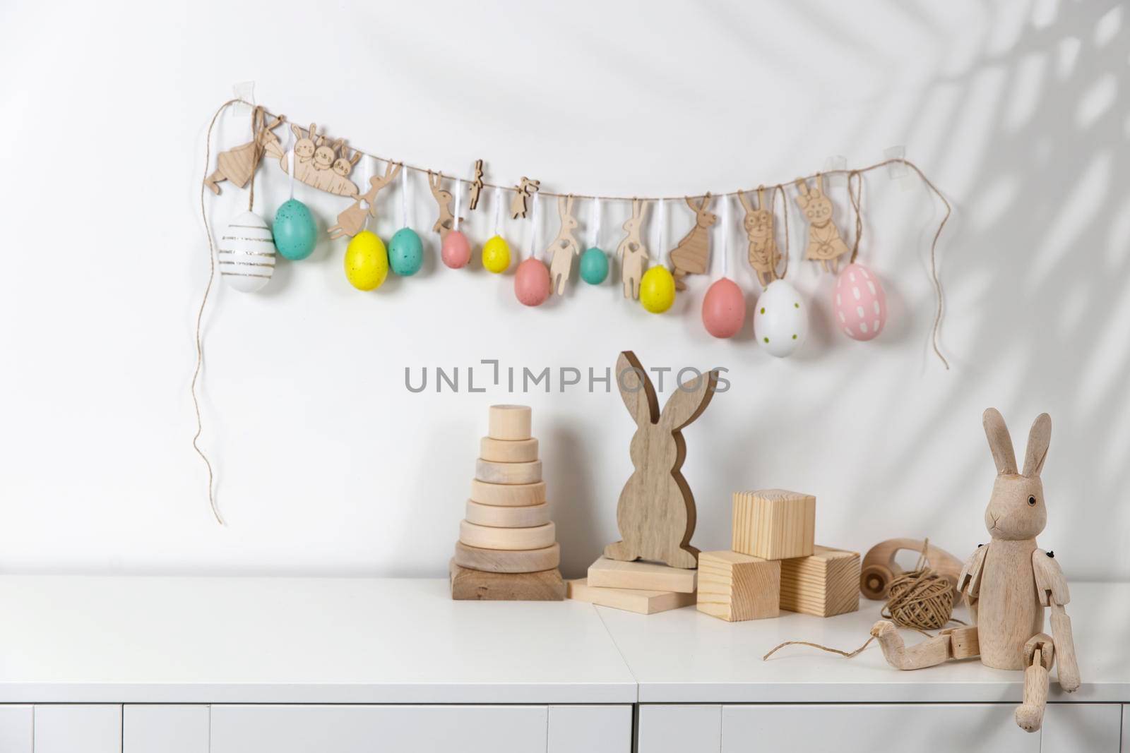 Fragment of the interior. Decorated children's room for Easter. A garland of plastic eggs and hares cut out of cardboard on the wall. Wooden rabbits and wooden cubes on the table. Place for your text. Easter card. by elenarostunova