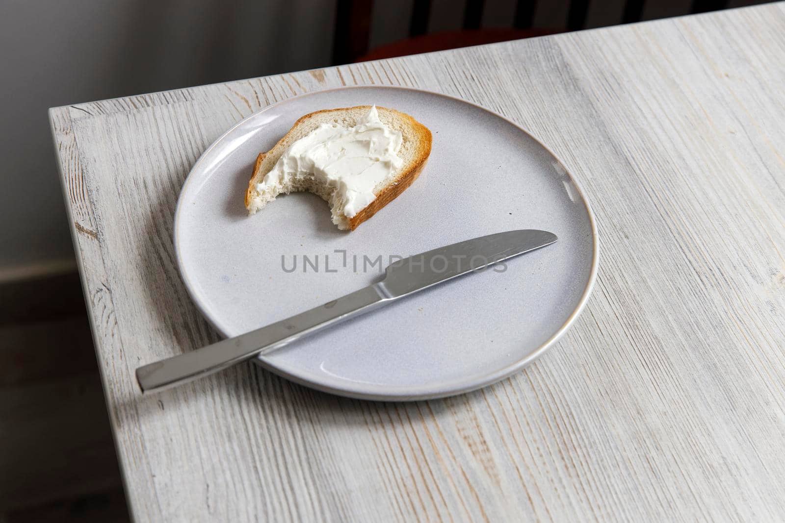 A piece of bread with curd cheese spread on it on a white ceramic plate with a knife on the beige table.