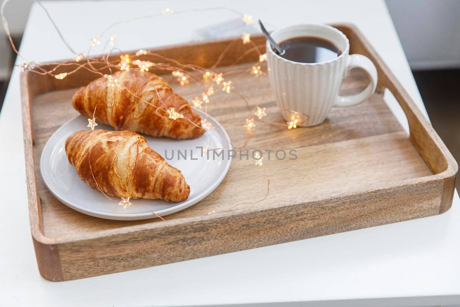 Freshly baked croissant on a gray round plate, white cup with coffee and garland on a tray on the table by elenarostunova