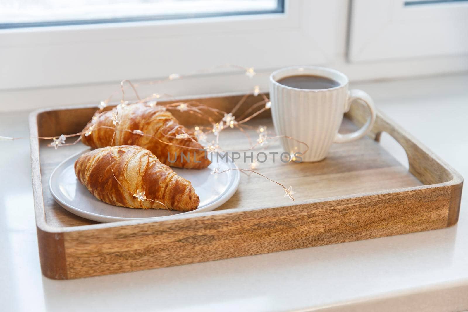 Freshly baked croissant on a gray round plate, white cup with coffee and garland on a tray on the table