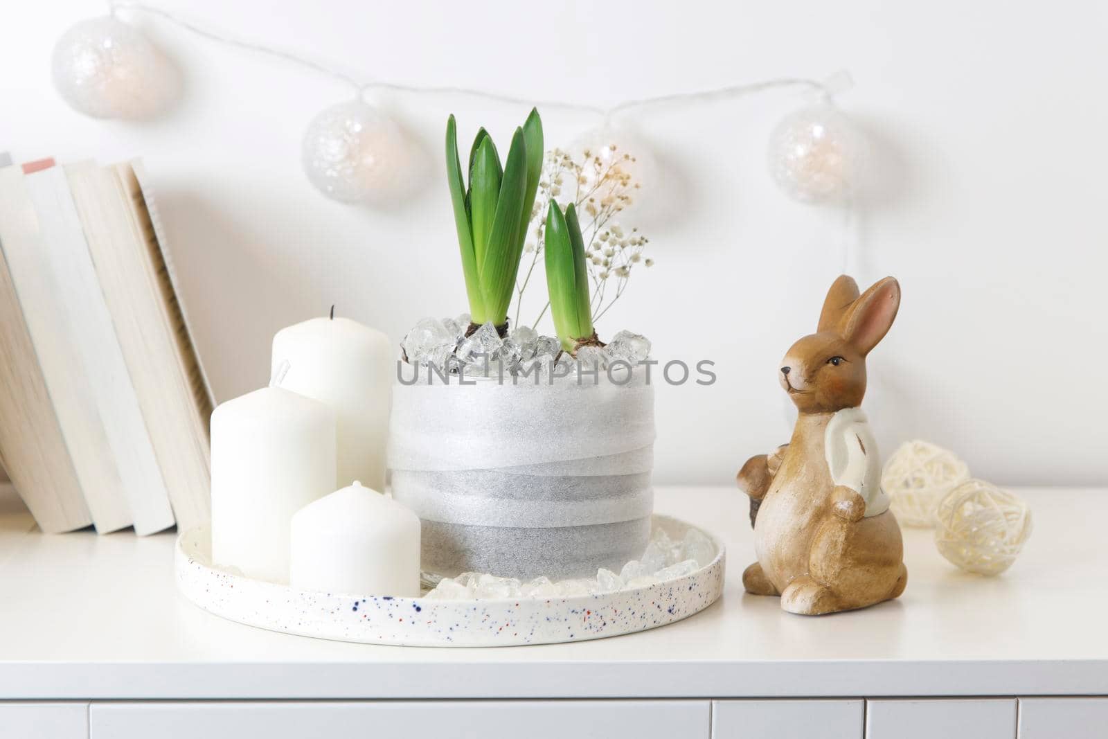 Unblown hyacinths with burning candles on a tray, a stack of books, a mug of tea, a garland of thread on the wall. Easter room decoration.