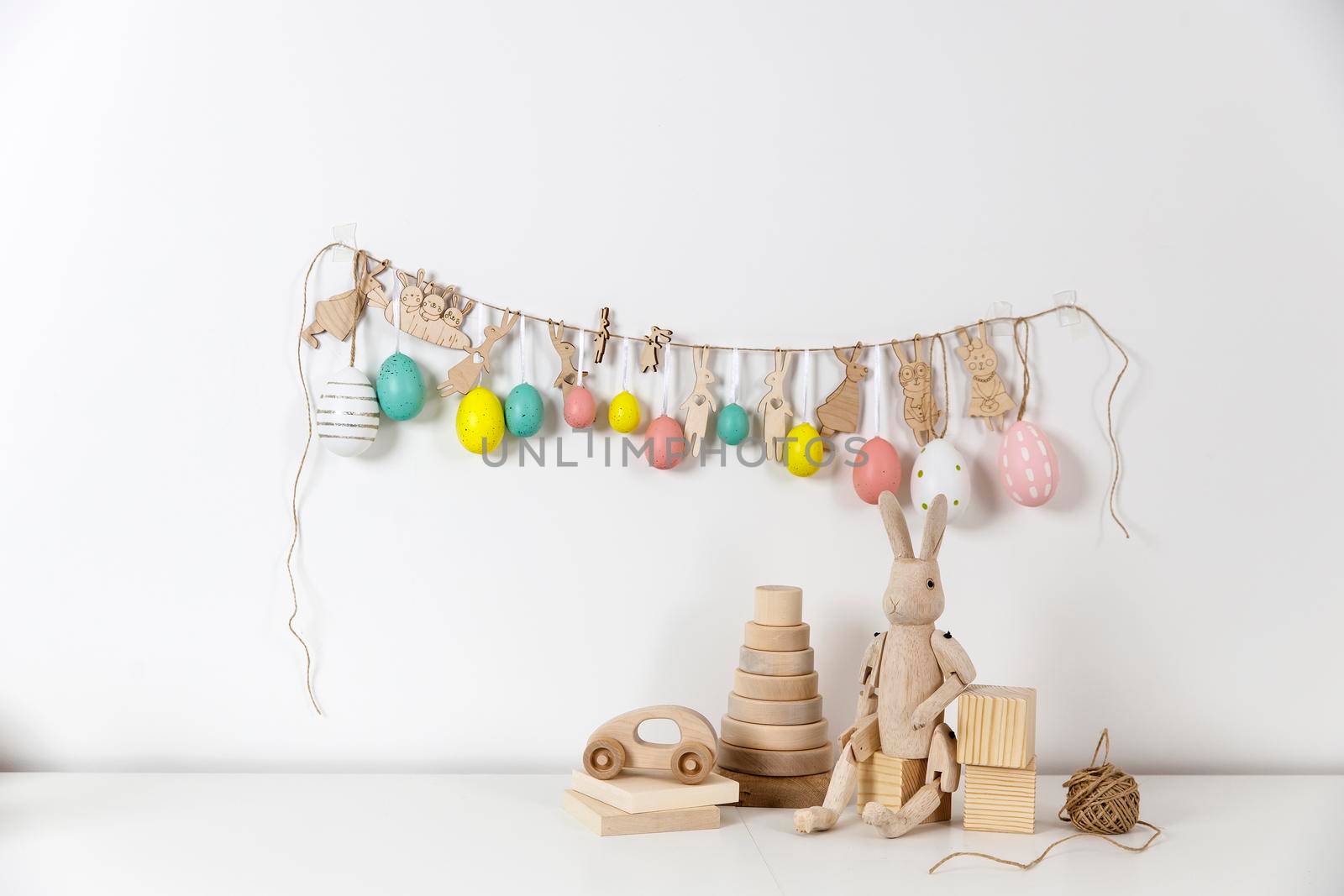 Fragment of the interior. Decorated children's room for Easter. A garland of plastic eggs and hares cut out of cardboard on the wall. Wooden rabbits and wooden cubes on the table. Easter card.