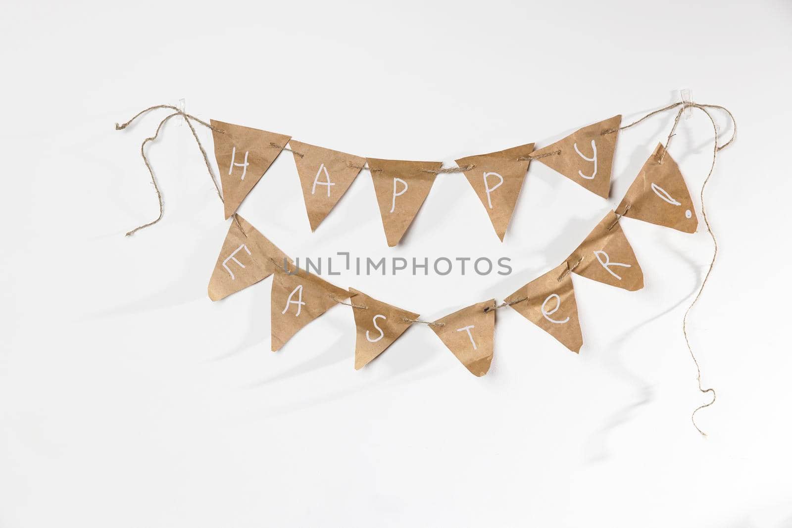 Homemade garland of craft paper flags with the inscription - Happy Easter hanging on a white wall by elenarostunova