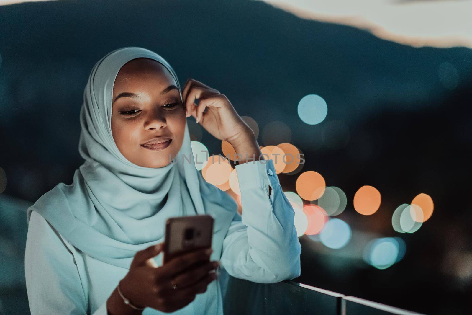 Young Muslim woman wearing scarf veil on urban city street at night texting on a smartphone with bokeh city light in the background. High-quality photo
