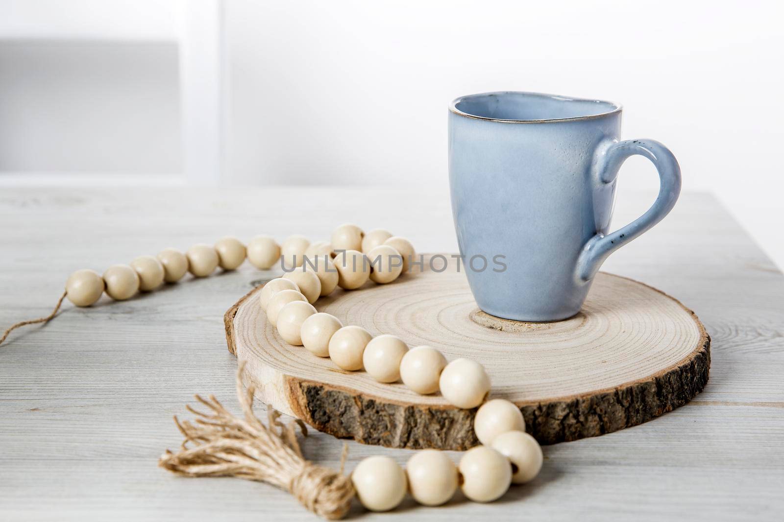 A blue ceramic cup on a wooden round cut, beads, a rosary for prayer on a beige table against a white wall. Place for text by elenarostunova