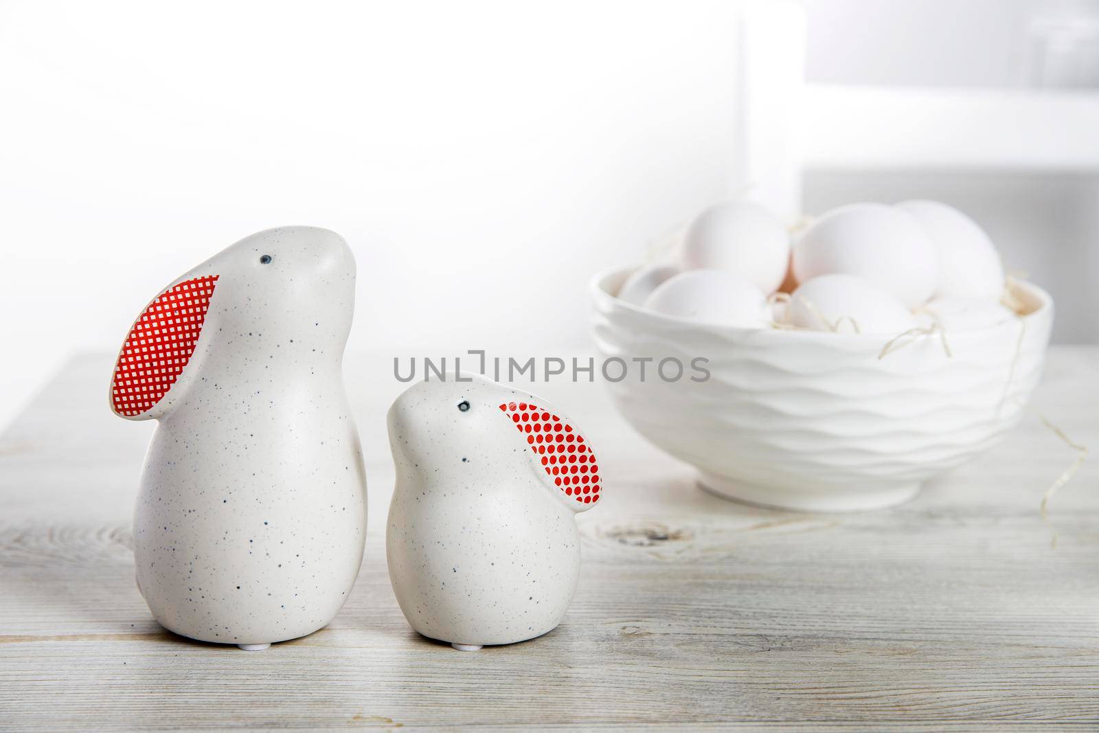 Two figurines of white hares with red ears big and small on a wooden saw cut and a bowl with eggs in the background in a white Scandinavian style kitchen by elenarostunova
