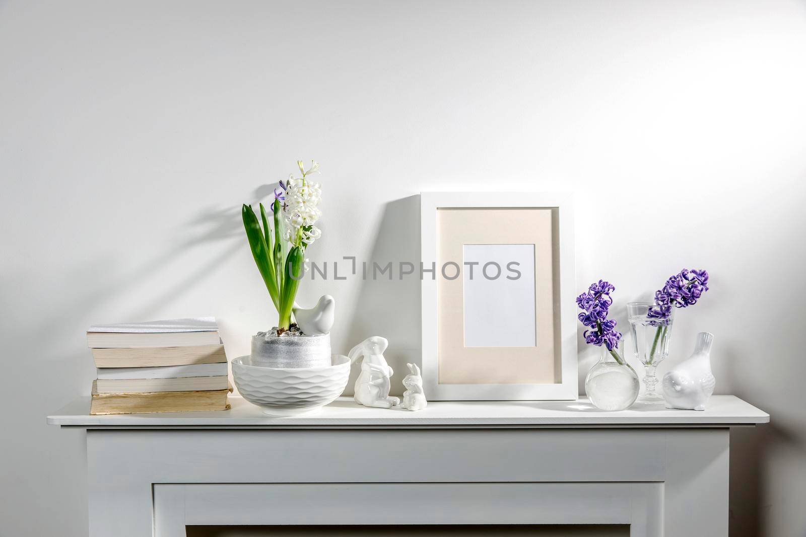 White hyacinth in a large porcelain bowl, books, figurines of hares and a bird, an empty photo frame are on the fireplace against the white wall. Layout.