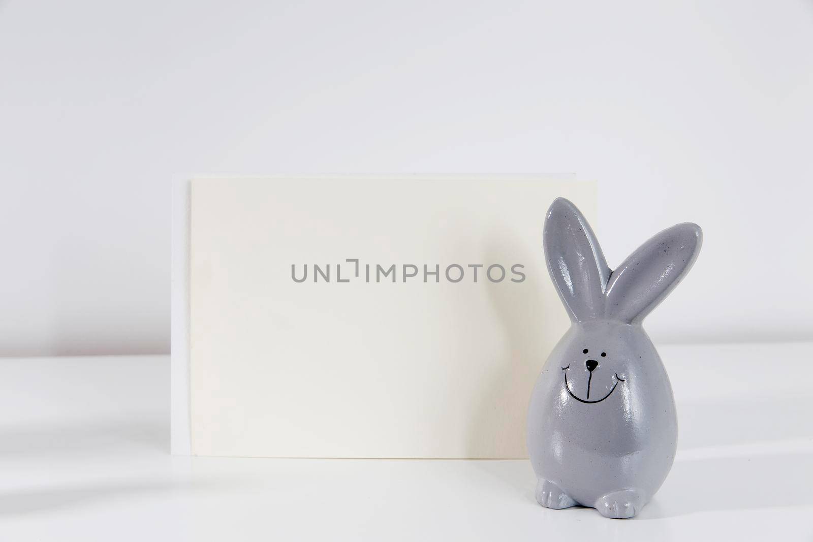 Mock up white frame with modern ceramic easter bunny decor on a shelf. White color scheme. Landscape frame orientation. by elenarostunova