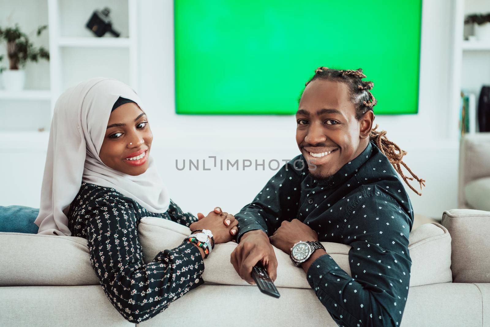 African Couple Sitting On Sofa Watching TV Together Chroma Green Screen Woman Wearing Islamic Hijab Clothes by dotshock