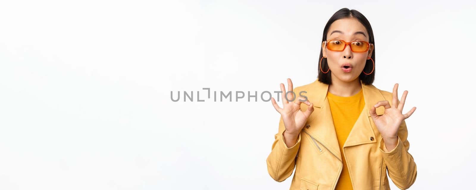 Enthusiastic asian girl in sunglasses, shows okay, ok sign in approval, smiling and laughing, approve, recommend smth, praise and compliment, standing over white background.