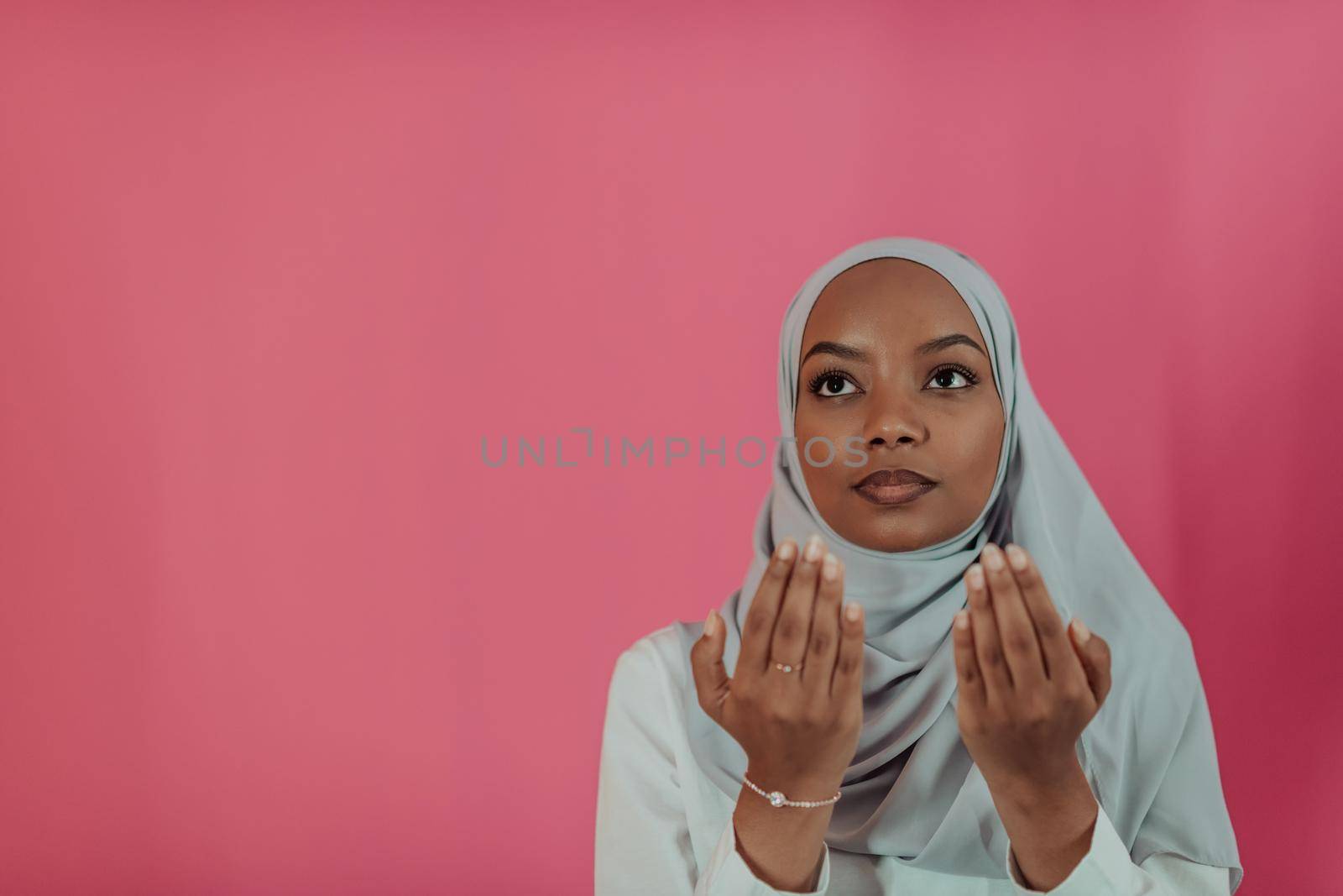 Modern African Muslim woman makes traditional prayer to God, keeps hands in praying gesture, wears traditional white clothes, has serious facial expression, isolated over plastic pink background by dotshock