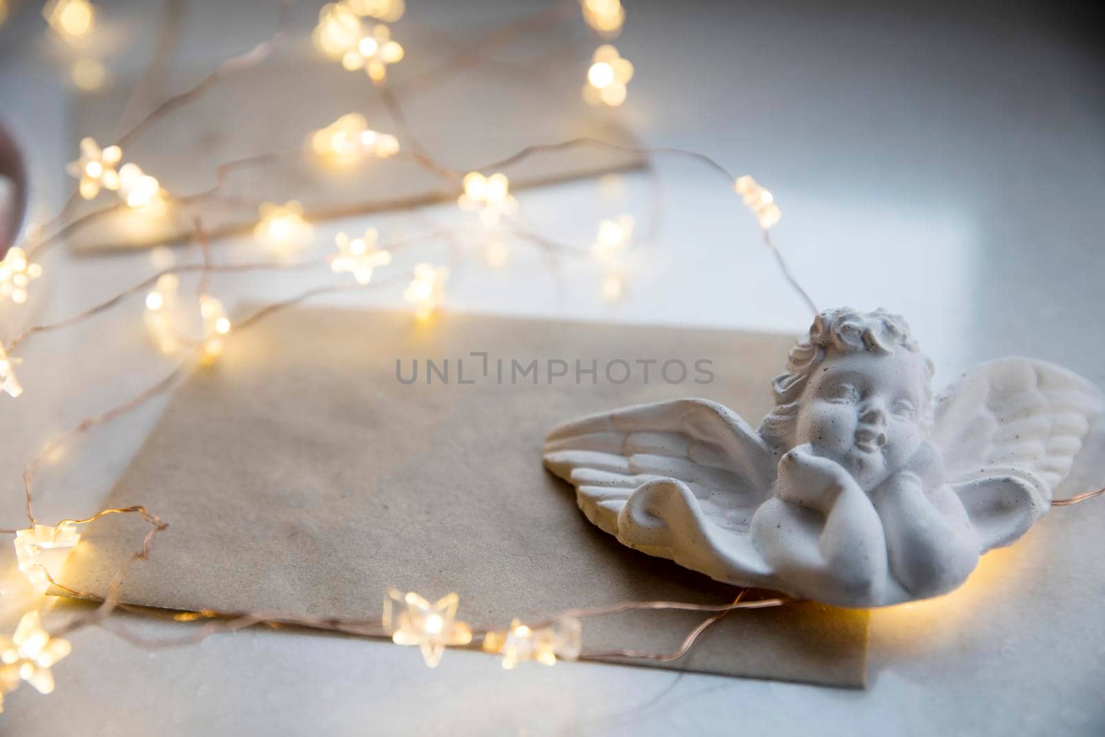 Apartment decoration for Christmas. A plaster figure of an angel is on a beige envelope with congratulations. An asterisk garland shines all over the frame. Copy space by elenarostunova