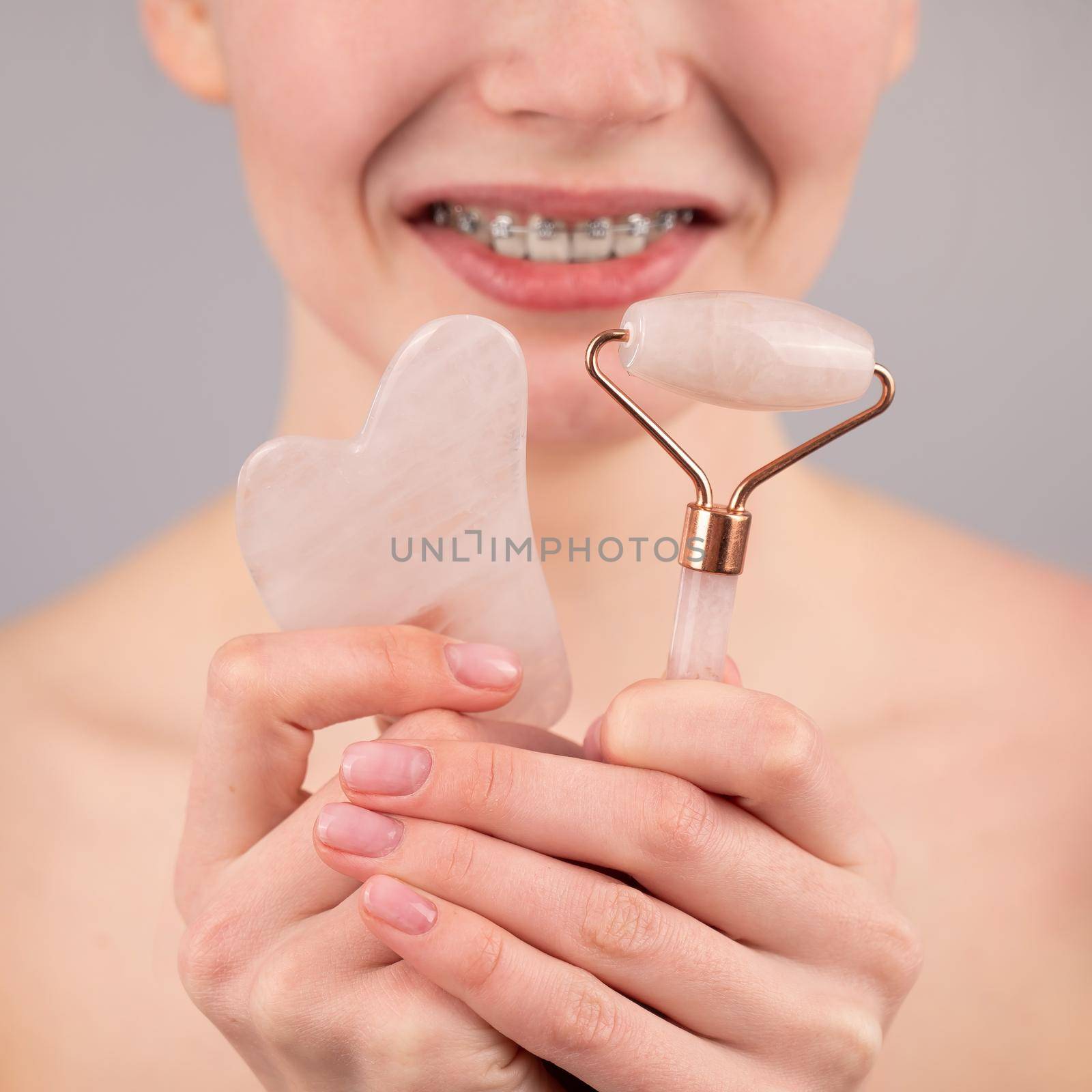 Close-up portrait of a woman with braces on her teeth holding a pink roller massager and a gouache scraper on a white background